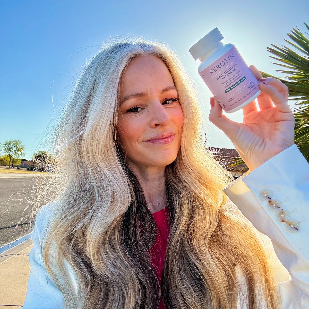 A woman with long blonde hair holds up a bottle of Kerotin Hair Growth Vitamins in her left hand, set against a bright outdoor background with blue sky and palm leaves.